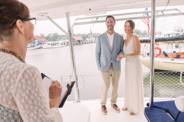 Couple on a boat getting married by an officiant, waterfront backdrop.