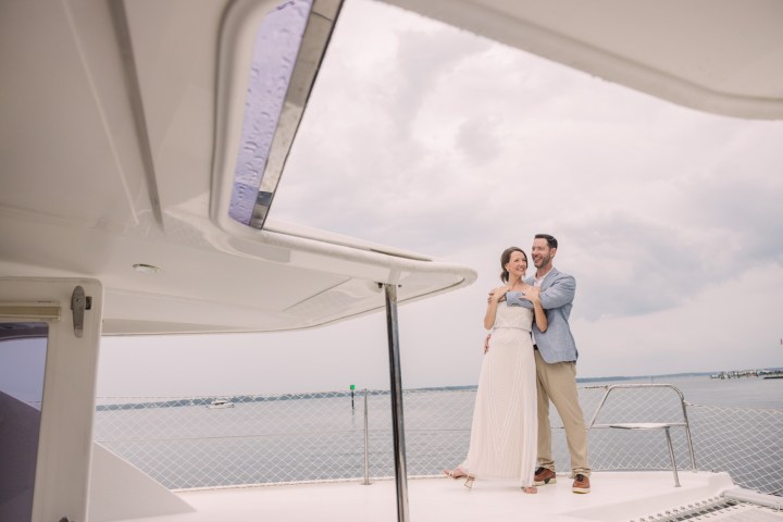 Couple smiling and standing on a boat deck with a cloudy sky and calm water in the background.