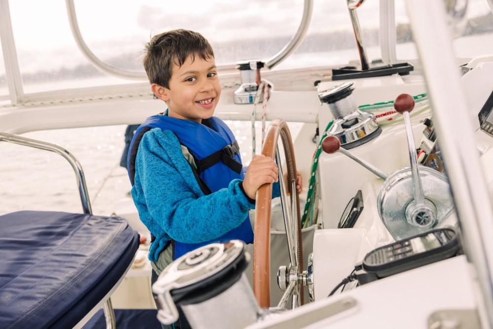 Smiling boy in a blue jacket steering a boat's wheel on a sunny day.