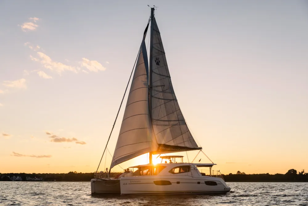 Sailboat on water at sunset with sails up and orange sky.