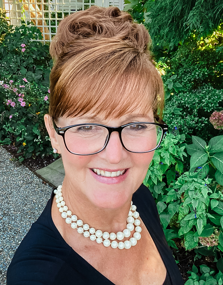 Smiling person with glasses and pearl necklace in a garden setting.