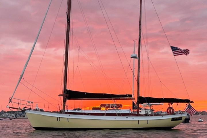 Sailboat on water with American flag, sunset in background with pink and orange sky.