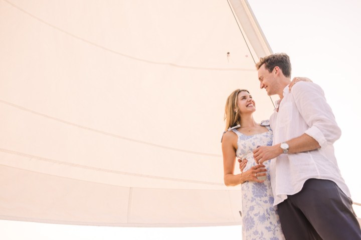 Couple smiling on a sailboat with large sail in the background.