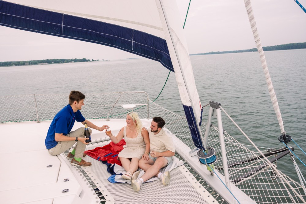 Three people relaxing on a sailboat, one pouring wine for a couple seated under a large sail.