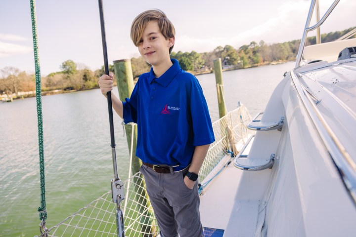 Young person in blue shirt standing on a boat by the water.