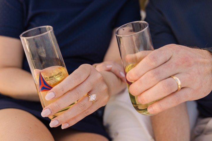 Two hands holding beer glasses, showing engagement and wedding rings.