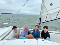 Four people sitting on a sailboat with cushions, smiling, against a cloudy sea backdrop.