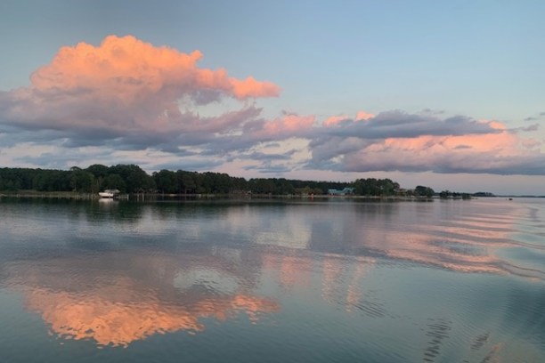 Pink clouds reflected on calm lake water at sunset, with trees lining the shore.