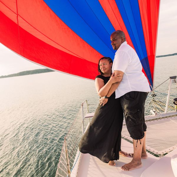 Couple embracing on a boat with a red and blue sail over water.