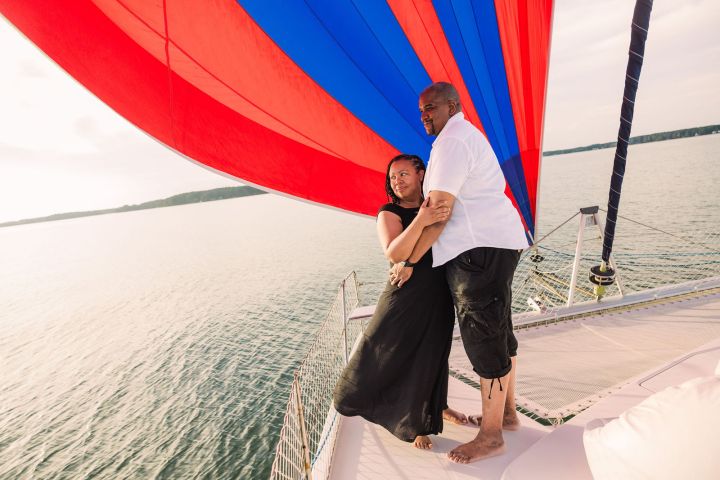 Couple embracing on a boat with a red and blue sail over water.