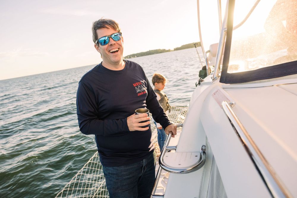Man smiling on a boat holding a drink, child in background, sunny day on the water.