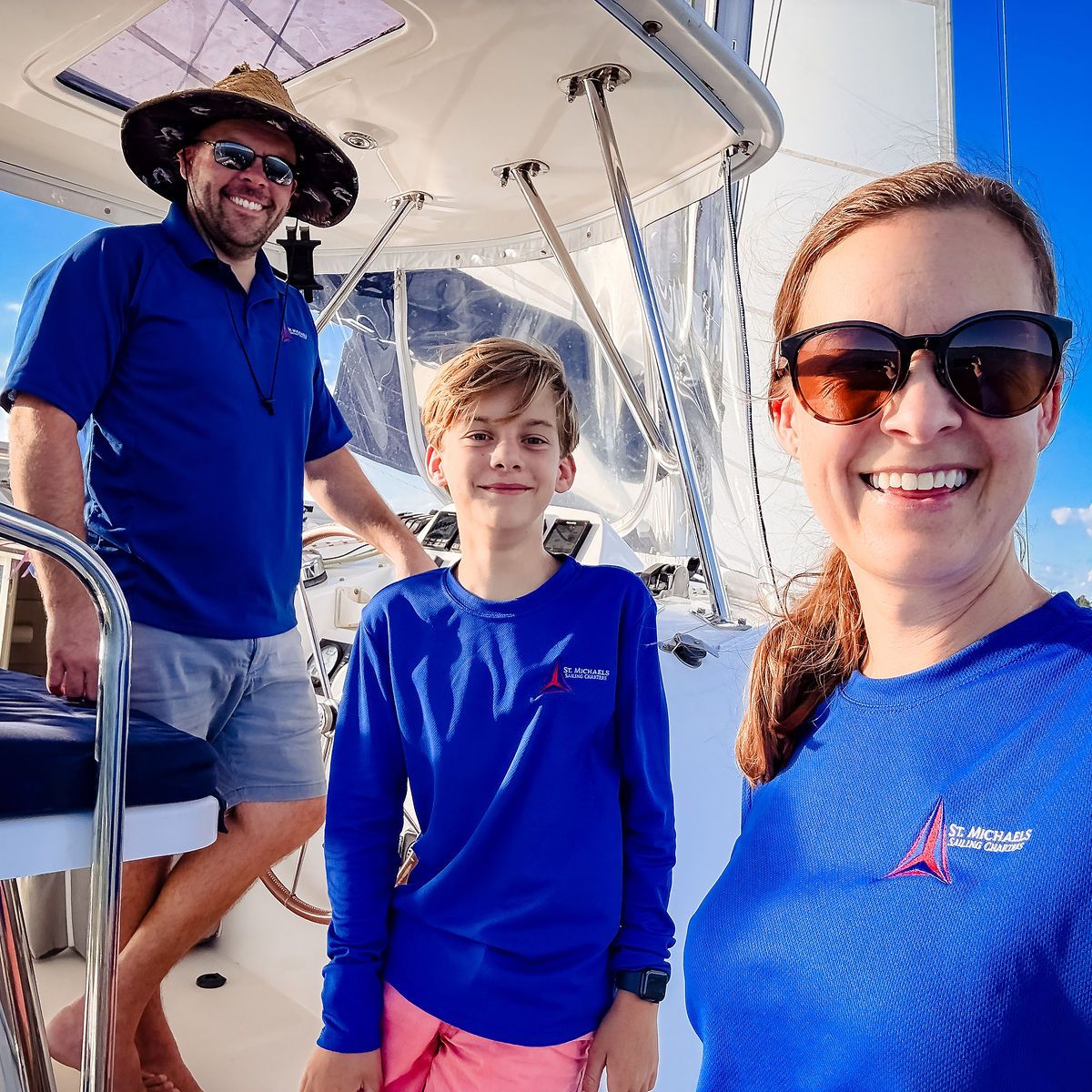 Three people in blue shirts smiling on a sailboat under clear skies.