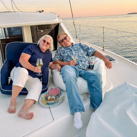 Two people relaxing on a boat, holding drinks, at sunset on calm water.