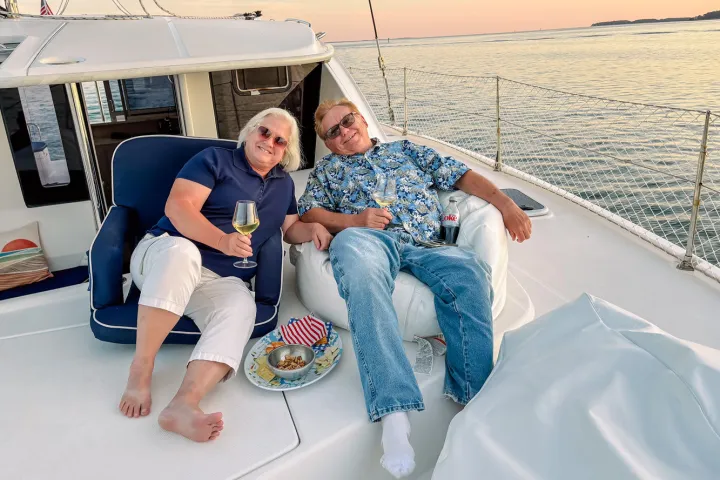 Two people relaxing on a boat, holding drinks, at sunset on calm water.