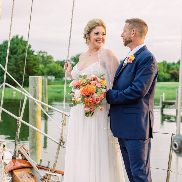 Bride and groom smiling on a sailboat with flowers, surrounded by water and greenery.