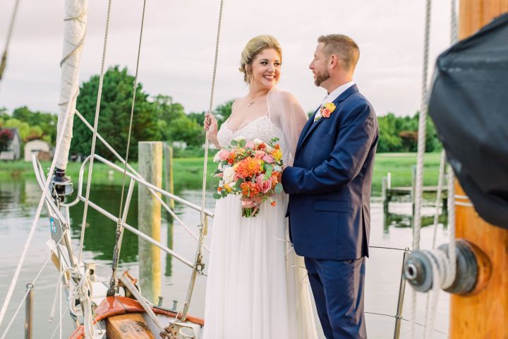Bride and groom smiling on a sailboat with flowers, surrounded by water and greenery.