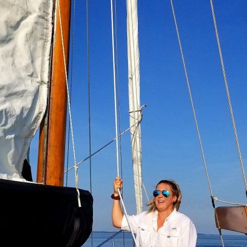 Person in sunglasses smiling on a sailboat, holding a rope against a clear blue sky.