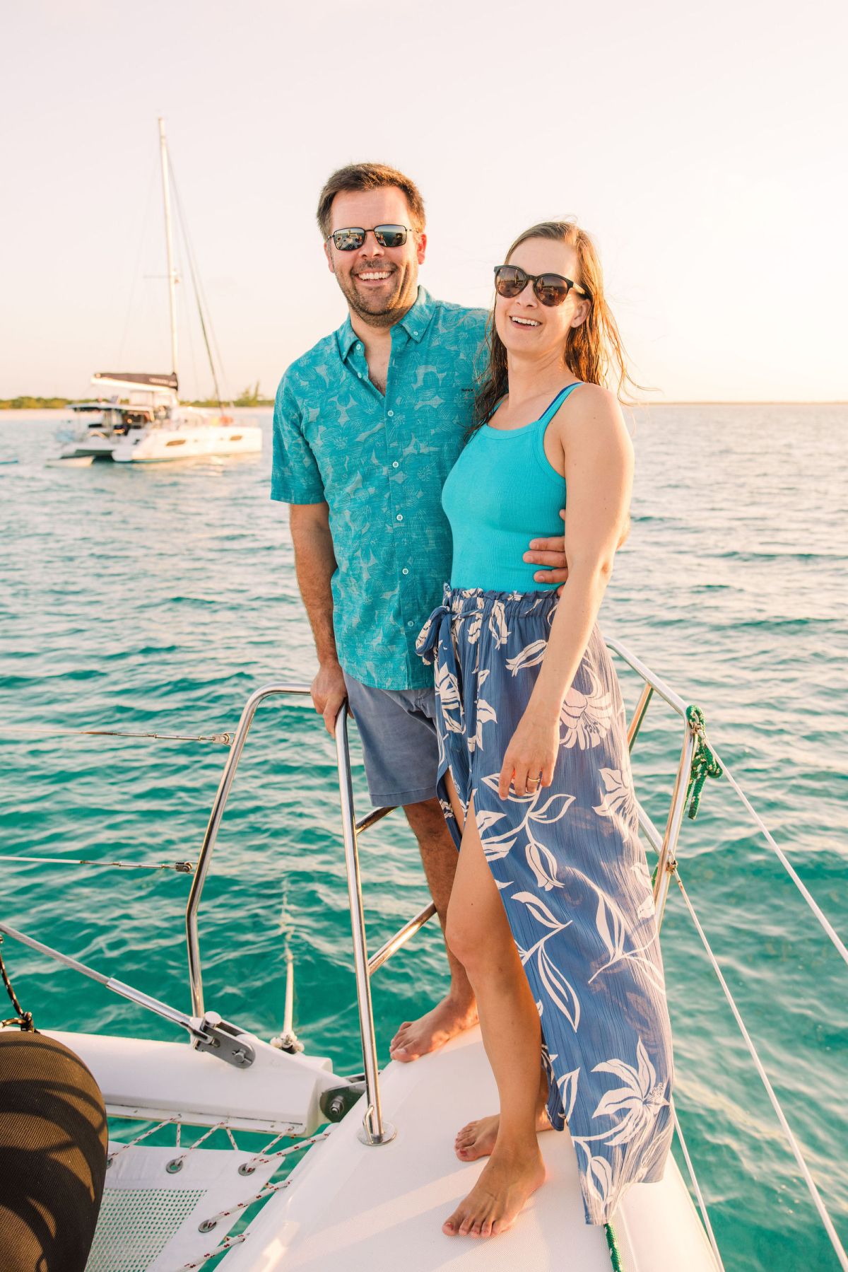 Smiling couple in sunglasses on a boat deck with ocean and sailboat in the background.