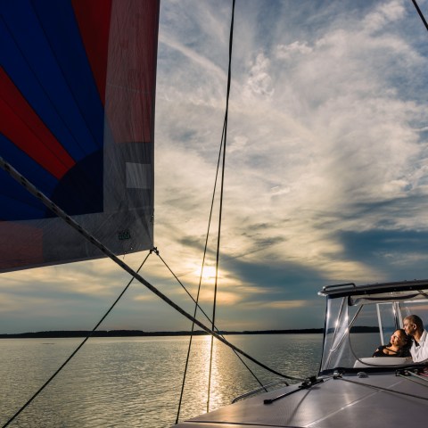 Couple enjoys sunset on sailboat with colorful sail on calm water.