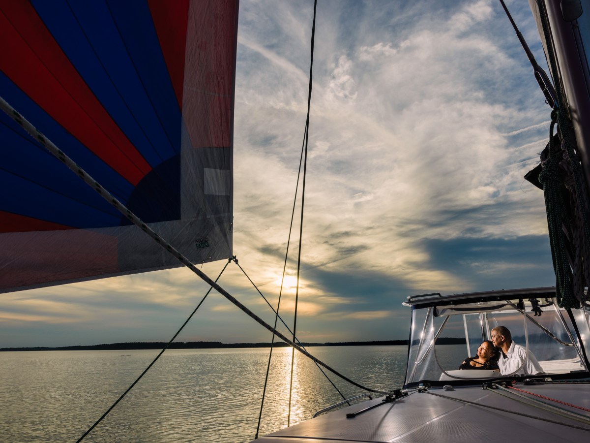 Couple enjoys sunset on sailboat with colorful sail on calm water.