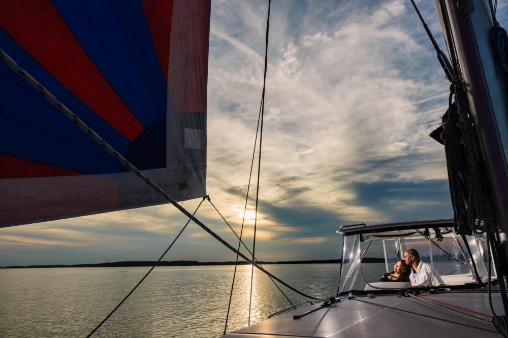 Couple enjoys sunset on sailboat with colorful sail on calm water.