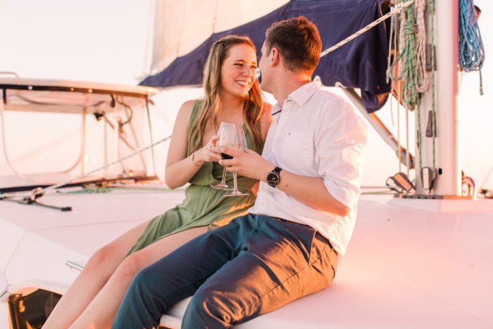 Couple sitting on a sailboat deck at sunset, holding wine glasses and smiling.