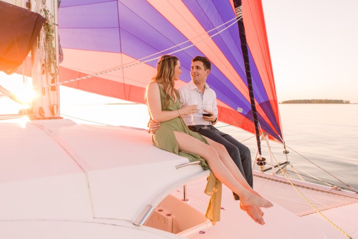 Couple sitting on a boat in sunset, holding glasses, with colorful sails in the background.