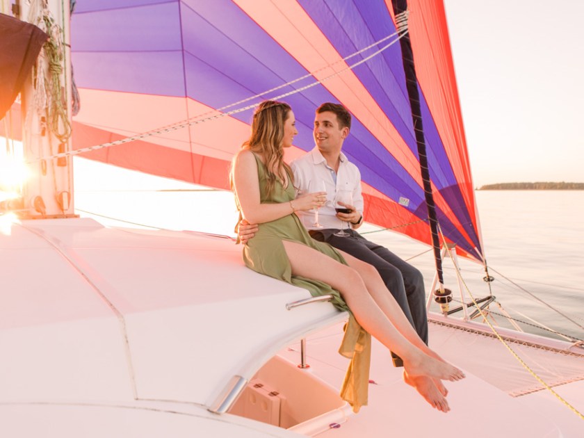 Couple sitting on a boat in sunset, holding glasses, with colorful sails in the background.