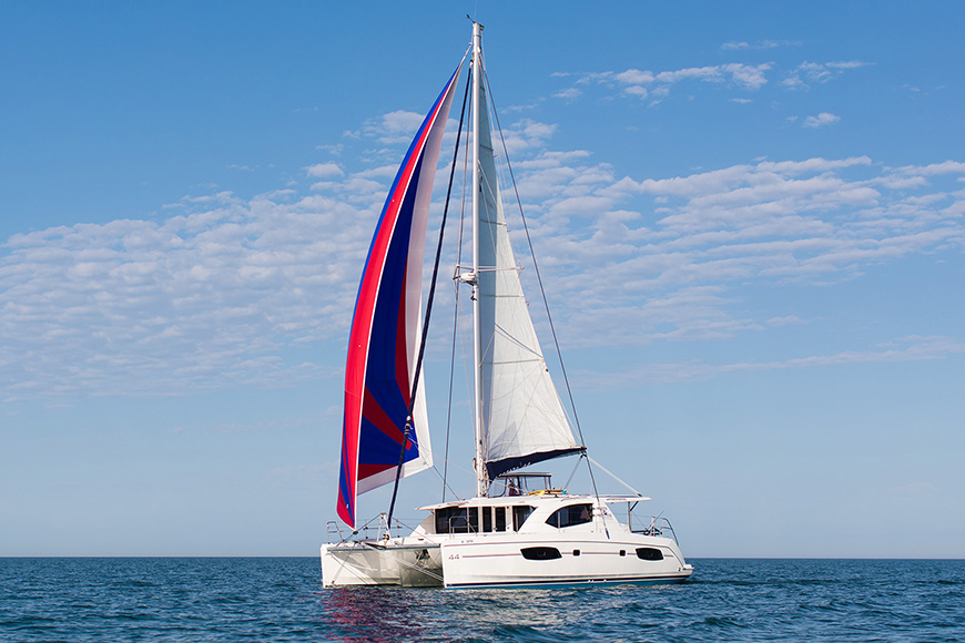 Catamaran with red and blue sails on open water under a blue sky.