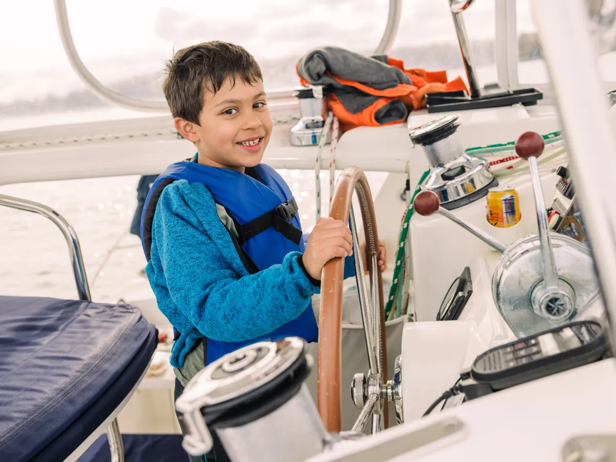 Smiling child wearing a life jacket steering a boat using a wooden wheel.