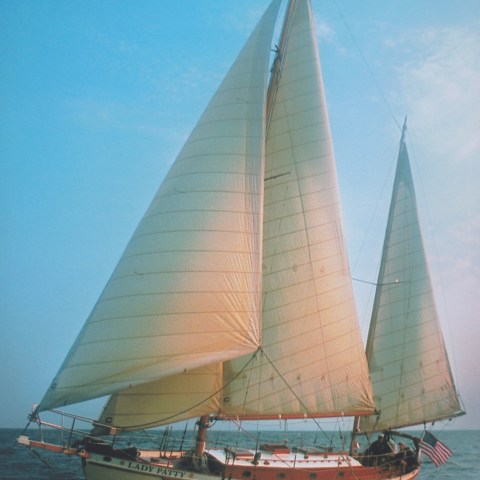 A sailboat with white sails on a clear day in open water, U.S. flag at stern.