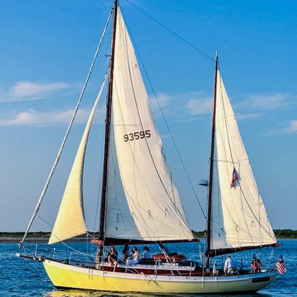 Yellow sailboat with three sails and people onboard in a sunny, blue sky setting.