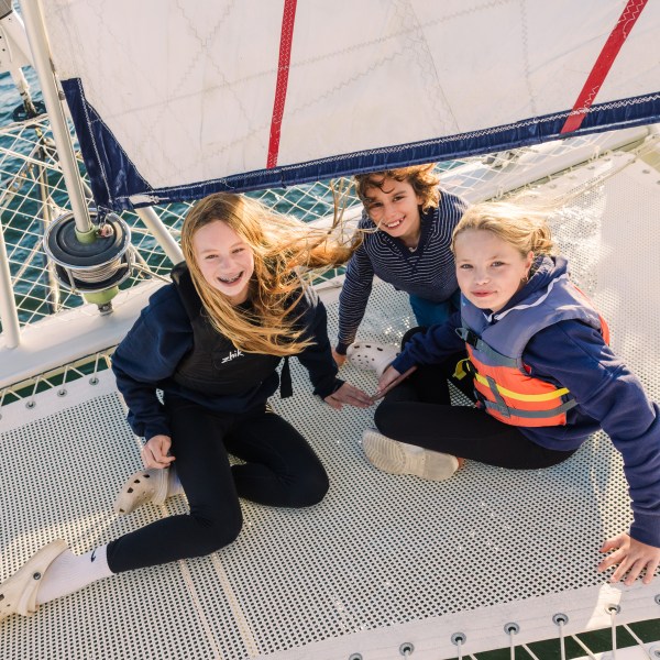 Three kids sitting on a sailboat deck with a sail above, smiling at the camera.