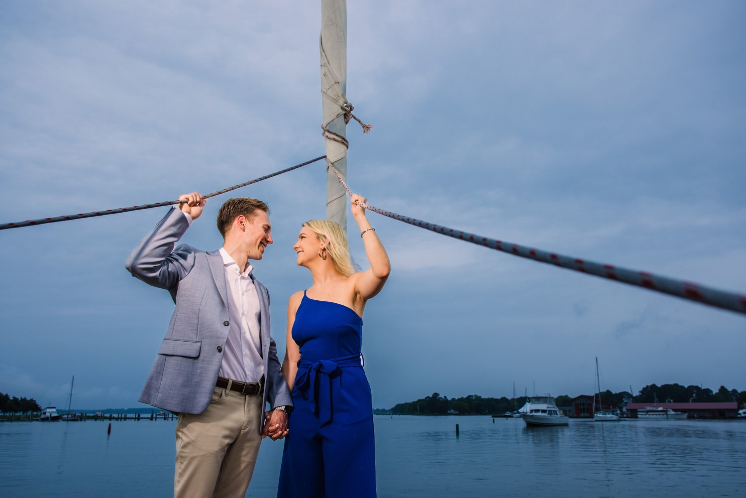 Couple holding a rope on a boat deck, smiling at each other, with a cloudy sky and ocean in the background.