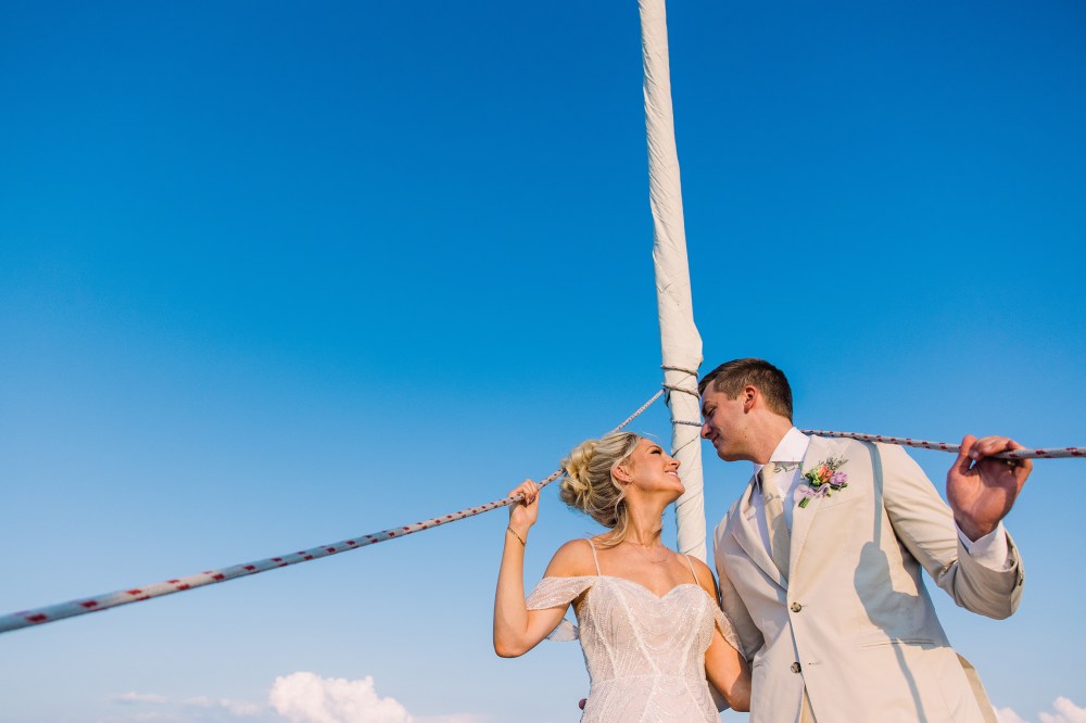 Bride and groom on a sailboat, holding ropes, under a clear blue sky.
