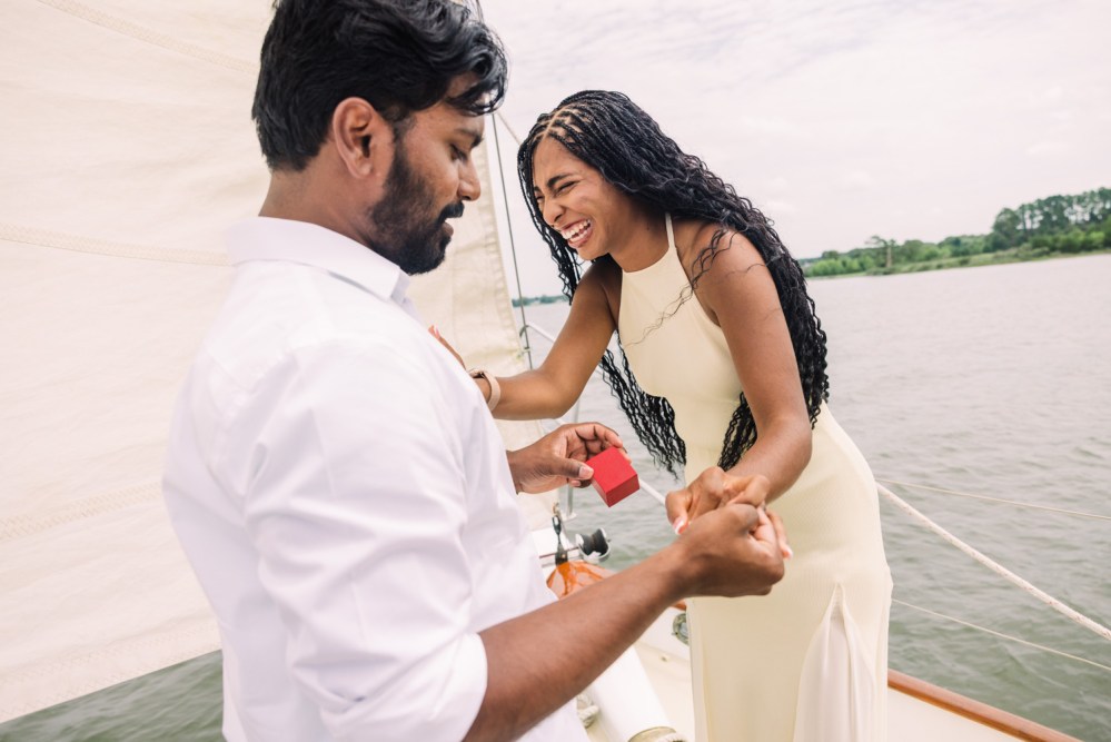 Couple on a boat; man holds a ring box, woman appears surprised.
