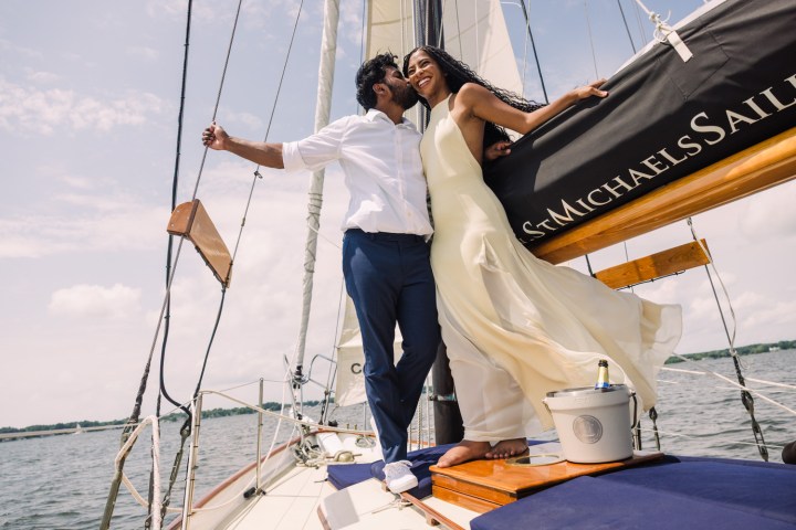 Couple smiling on a sailboat in sunlight, with sails behind them.