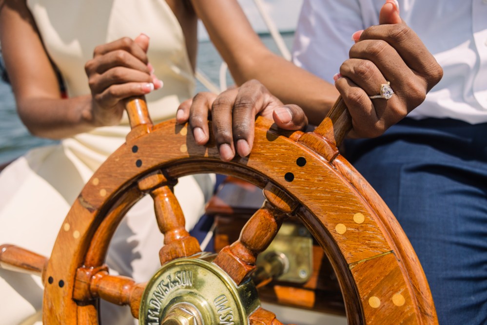 Two hands on a ship's wooden wheel, one with an engagement ring.