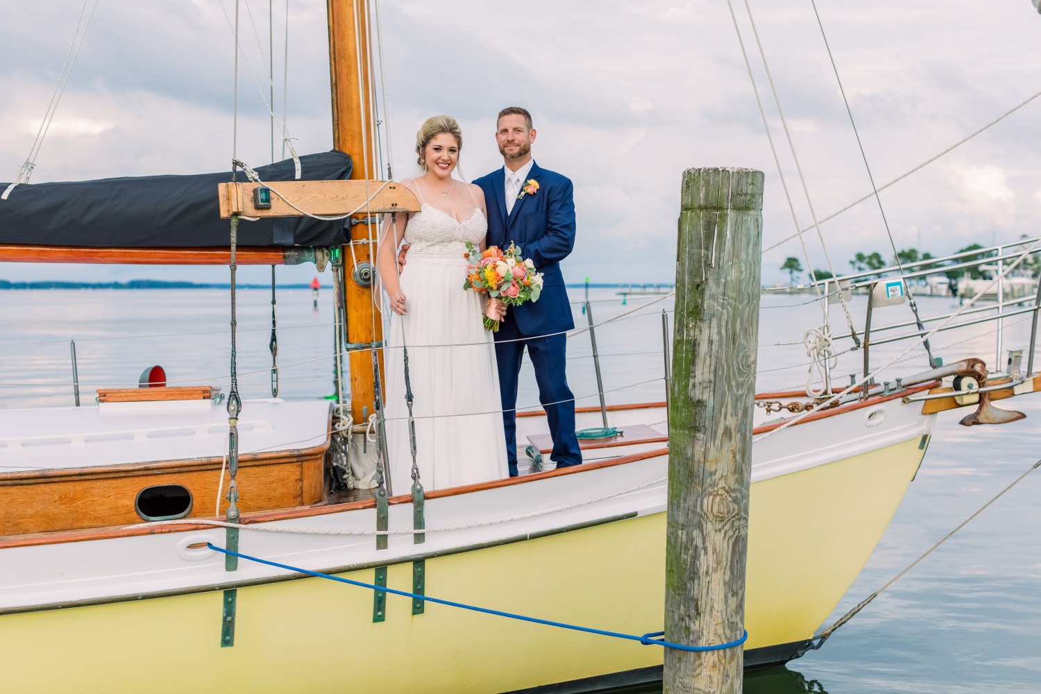 Bride and groom standing on a sailboat with a wooden mast and calm water in the background.