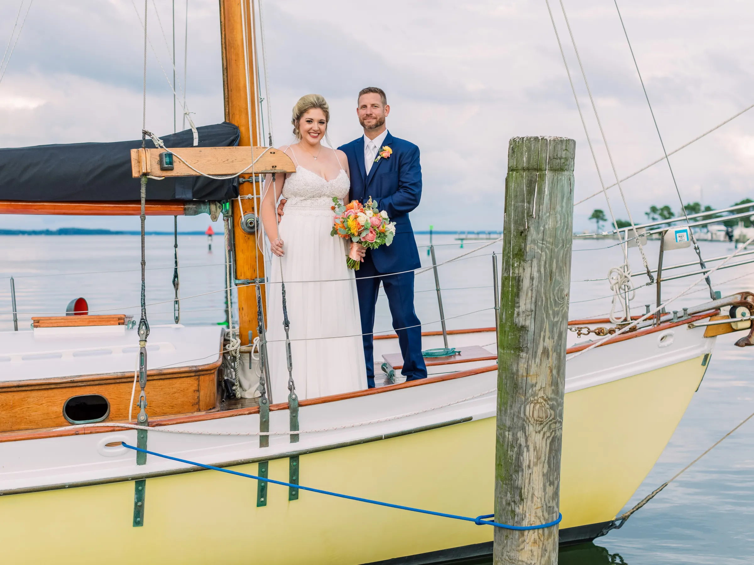 Bride and groom standing on a sailboat with a wooden mast and calm water in the background.