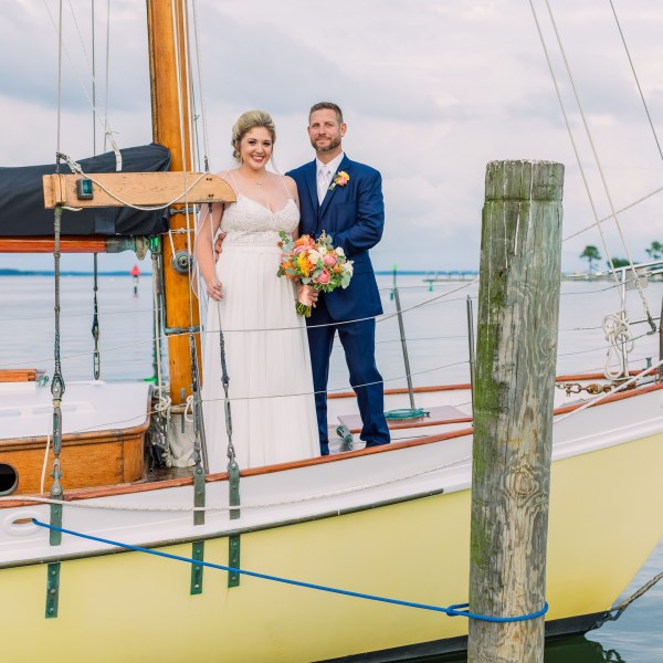 Bride and groom standing on a sailboat with a wooden mast and calm water in the background.