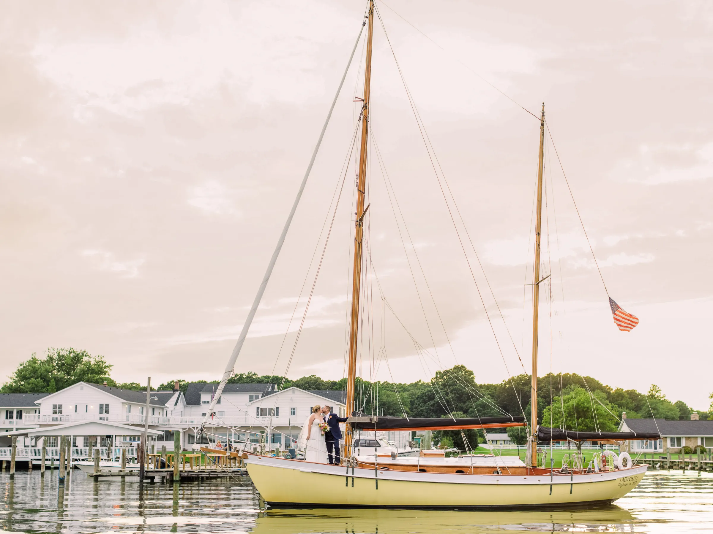 Couple on sailboat with American flag near dock, buildings in background.