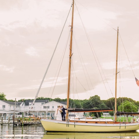 Couple on sailboat with American flag near dock, buildings in background.