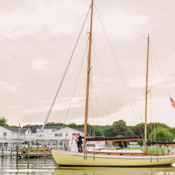 Couple on sailboat with American flag near dock, buildings in background.