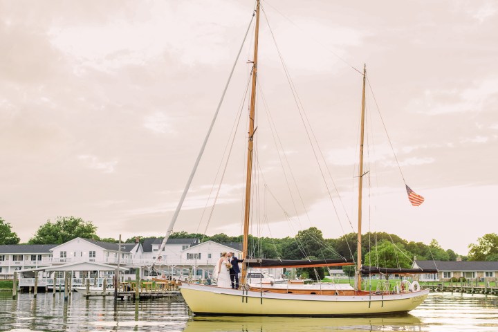 Couple on sailboat with American flag near dock, buildings in background.
