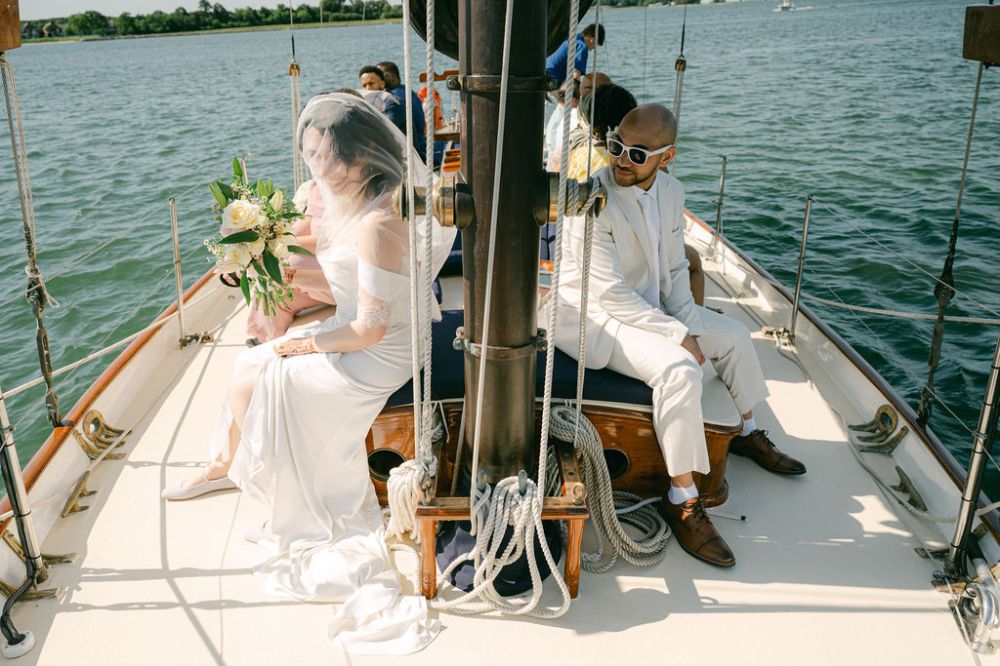 Bride and man in white attire sitting on a sailboat on a sunny day.