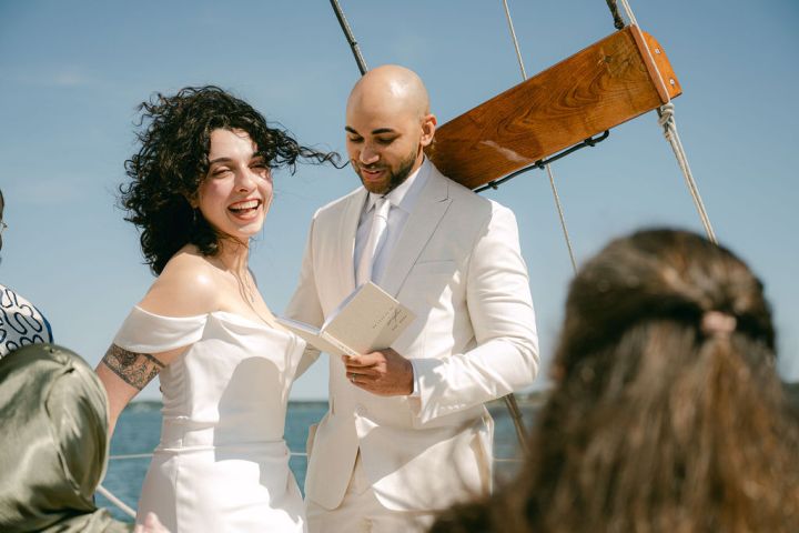 Couple in white attire smiling during outdoor wedding ceremony on a boat.