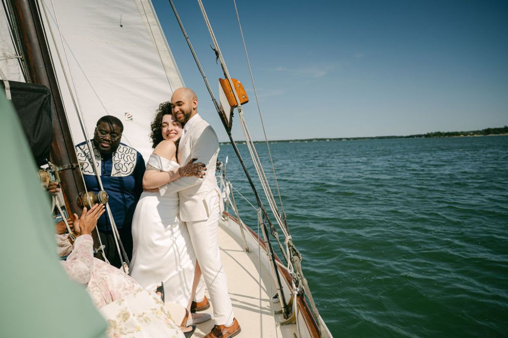 Bride and groom hugging on a sailboat with a cheerful friend nearby.