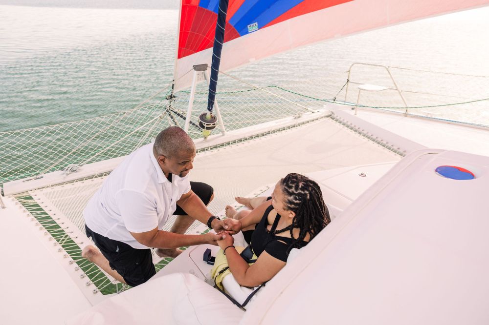 Man kneeling on a sailboat holding a woman's hands under a colorful sail.
