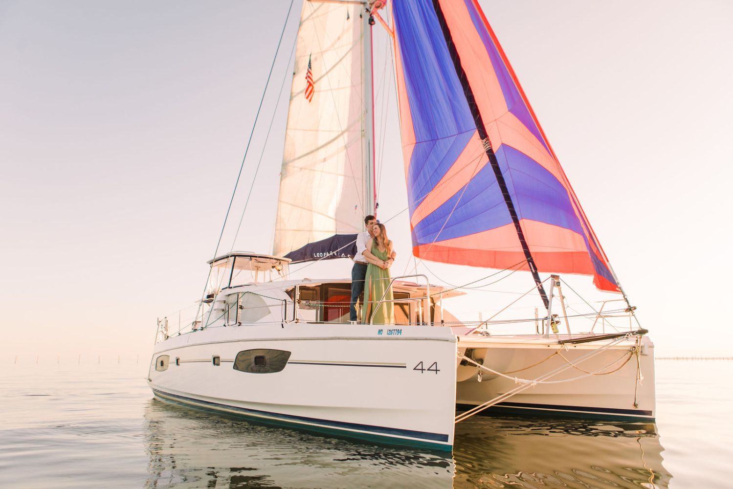 Couple standing on a sailing catamaran with colorful sails at sunset.